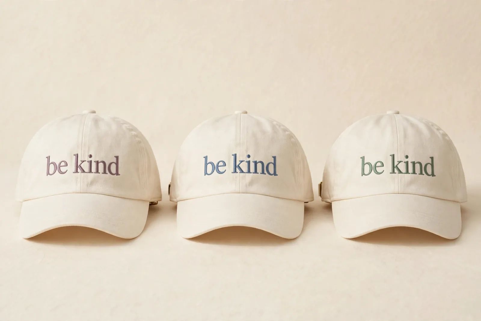 Three soft cream dad caps in a row, each embroidered with only the words be kind in serif lettering: mauve on the first, dusty blue on the second, sage on the third